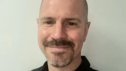 Ross Ayres has brown eyes, a shaven head, moustache and cropped goatee beard. He is wearing a black top.
