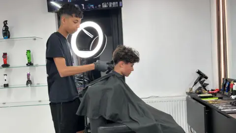 A boy is sitting on a barber's chair with a black gown on. Another boy is cutting his hair and is wearing black gloves.