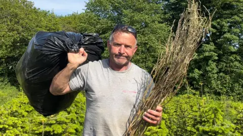 Rob Morgan is holding a black bin bag over his left shoulder and a garden waste in his right hand. He is wearning a grey t-shirt and has black sunglasses on top of his head. He is not smiling, with a fed up expression. 