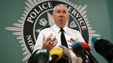 Getty Images Chief Constable Jon Boutcher stands in front of several media microphones wearing a white shirt with black PSNI badges and a black tie. A large PSNI logo looms over him in the background.