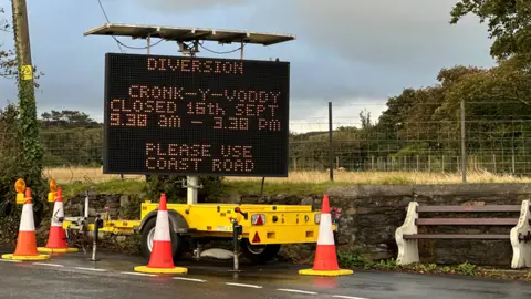 A mobile matrix sign on a yellow trailer directing motorist to use a diversion via the Peel Coat Road because of the road closure at Cronk-y-Voddy between 09:30 and 15:30 on 16 September. The trailer is surrounded by orange and white traffic cones and there is a public bench to the right of it with fencing, grass and trees behind.