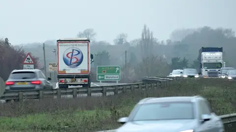 DJ McLaren/BBC Another view of the A1 dual carriageway on a cloudy and grey day. Vehicles travel either side of the road. There is a green road sign toward the middle of the image.