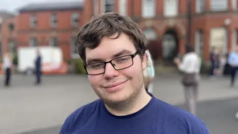 A man with short brown hair and glasses looks at the camera. He wears a navy t-shirt and has short stubble on his lower chin. Behind him is an out-of-focus red-brick building.