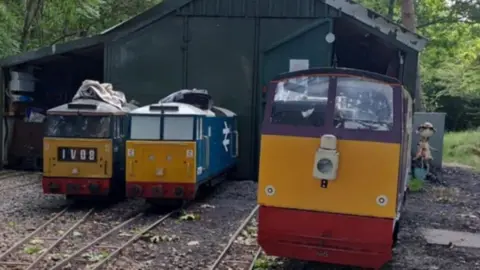 Bob Brocklehurst This photograph shows a small railway shed housing miniature locomotives.
Dense green foliage and trees are visible in the background.
The shed is made of corrugated metal panels painted dark green, with a pitched roof. Multiple narrow-gauge railway tracks lead into the shed.
Three miniature locomotives painted red, yellow and blue are positioned on separate tracks in front of and partially inside the shed. 