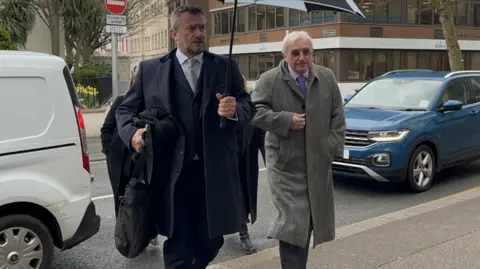 Two men in a street, one holding an umbrella andn another in a long coat.