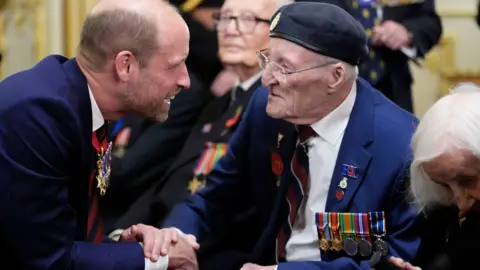 An elderly man in a blue suit, wearing military medals, sits in a wheelchair as he is shakes hands with Prince William. Other people can be seen sat beside him.