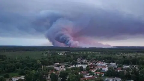 A drone view showing the Sainte-Helene commune in France with smoke rising in the background during a wildfire in a forest area in Saumos
