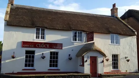 The Clock House White thatched pub with dark red signage