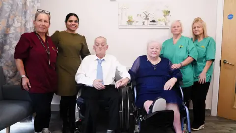 Colin and Pamela sat next to each other in their wheelchairs in the care home with the staff standing next to them. On the left, two women are stood next to each other. One is wearing red nursing uniform. The other woman is wearing a green dress. On the right, next to Pam, two women are wearing green nursing scrubs. 