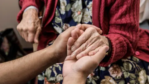 A close-up of two people holding hands. One of the hands belongs to an older person, who wears a floral dress and red cardigan.