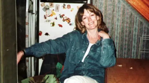 BBC Arlene Fraser sitting with her left elbow resting on a wooden table. She is wearing a blue denim shirt, white trousers and a white top. Behind her can be seen a fridge with a number of fridge magnets, patterned wallpaper and a wooden beam.