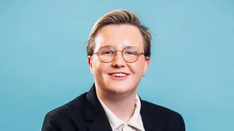 Handout Oscar Brooker smiles, wearing glasses against a blue background