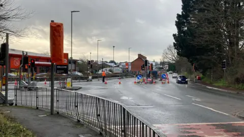 Roadworks and temporary traffic lights in front of a zebra crossing. There's a house in the background with road work barriers and cones in the foreground. There's a B&Q shop and queues of cars.