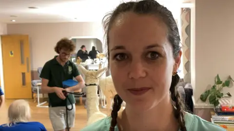 Luke Deal/BBC Magdalen House Care Home's activities manager Marianne Refurn with an alpaca and its handler inside the home in the background