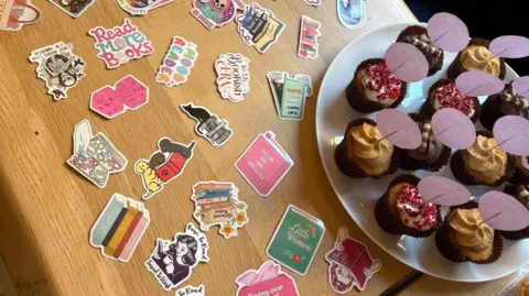 Lauren Emms Book-themed stickers spread out on a wooden table. There is a plate of cupcakes with gold icing. 