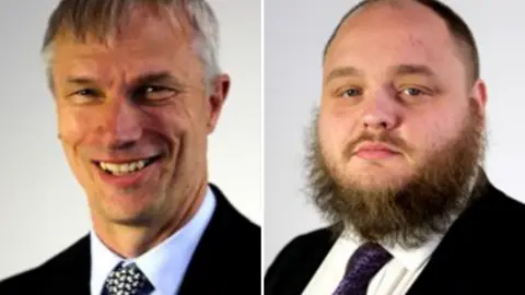 A side by side shot of two men in suits. The man on the left has short grey hair and the man on the right has a skin fade with a short unruly black beard