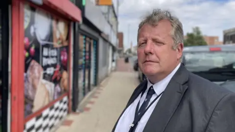 A man with short grey hair is pulling a serious expression for a landscape head and shoulders shot and looking into the camera whilst standing on his side. He is wearing a grey blazer and white shirt with black tie with white dots on it. Behind him is a blurred corner shop and a street stretching out.