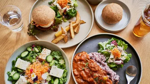Aerial view of four plates of food. One is a salad, another burger and chips, another is a rise dish with curry and the final one is a bun. There is a glass of water and beer also on the wooden table.