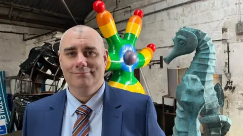 Andrew Turner/BBC Daniel Candon has a close cropped head of hair. He is wearing a blue suit, a light blue shirt and a red tie with light and dark blue, and white, stripes diagonally across the design. He is standing in a warehouse with several sculptures around him.