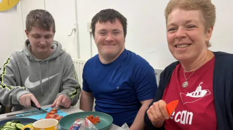 Two young men are cutting tomatoes for a pasta salad. Reece is actively cutting and Connor is sat in front of a plate of cut tomatoes smiling. Reece is wearing a light grey jumper with a big white nike tick on the front. Connor wears a dark blue t-shirt. Sheila is a volunteer and wears a red t-shirt with a submarine on it, she is holding an orange knife.