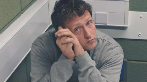 CCTV image of france in police custody. He is in a police interview room with grey brick walls. He is wearing a grey jumper and has curly brown hair. His elbows are resting on the table with his hands up against the left side of his face, He is looking directly at the camera.