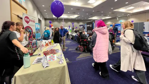 People walk around stalls and stands in a hotel lobby, There are signs and banners up with charities displaying information on their tables. 