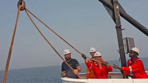 Four men in white hardhats, one in a navy T-shirt and the others in orange overalls hold ropes aboard the Léon Thévenin - the blue sea can be seen behind them