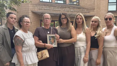 Seven woman stand together in a group. One is holding a framed photo of Darcy Ross. They all look at the camera.