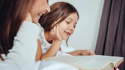 Getty Images A young girl with brown hair is lying down with an open book in front of her. A woman with brown hair is lying next to her and smiling and also looking at the book. In the background in a purple curtain.