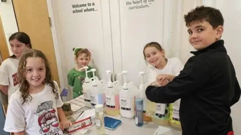 Five children running their eco-refill shop at a school. There plastic containers full of liquid on a table, ready to be pumped out.