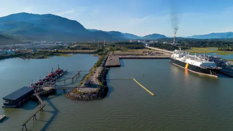 An image showing an LNG carrier loaded at the LNG Canada docks in Kitimat, British Columbia. There is port infrastructure visible, a large ship and LNG flares in the background emitting from the facilitiy. It is set on the water, with a picturesque backdrop of small mountains and pristine, green landscape.