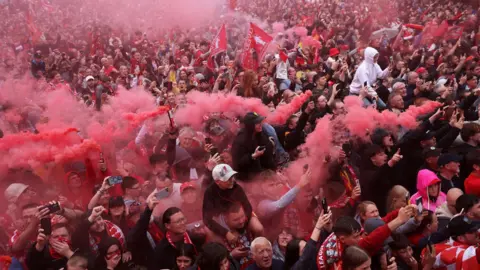 A large crowd of Liverpool fans are holding smoke bombs in a crowd as they watch the parade bus pass