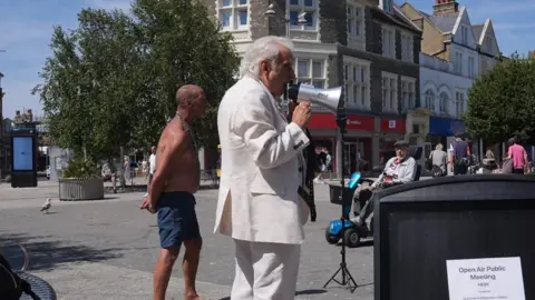 PA Media Erlam viste un traje blanco y habla por un megáfono.