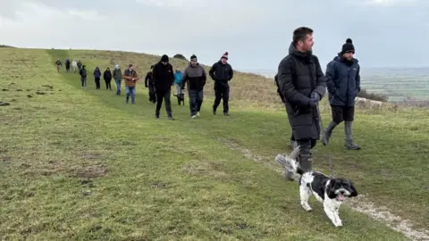 A line of men along with a couple of dogs, most dressed in anoraks and woollen hats, walking along a path on a grassy hillside with fields stretching off into the distance. The sky is cloudy.