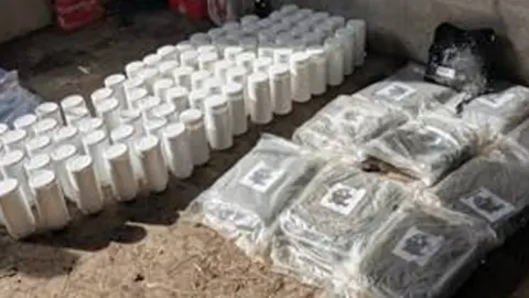 Items recovered by police officers from a rural shed in the Dunadry area of County Antrim. The image shows transparent bags full of a green substance and a series of white cannisters in rows.