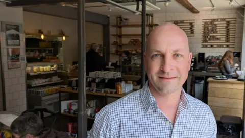 Anthony Harrison in checked shirt inside a bakery with loaves and till visible behind him