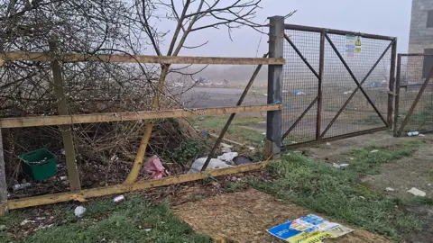 LDRS A wooden fence and metal gate at the entrance to the development. There is rubbish on the ground and derelict land behind 