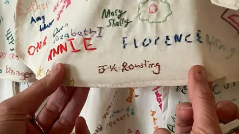 A pair of hands are holding a piece of fabric covered in embroided names. The one being held prominently to the camera is "JK Rowling"