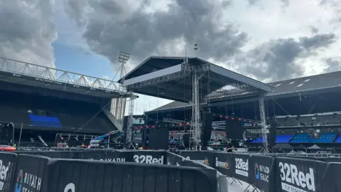 Dino Cabras Large covered stage in the centre of the image. There are two football stands in the background. The sky is blue with moody grey clouds coming in above the stadium.