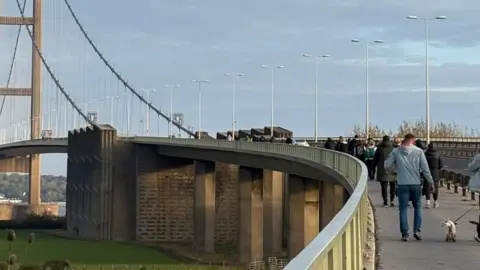 Gareth Morrison Rear view of a group of people walking over the Humber Bridge. It is a clear day and there are two dogs visible in the image