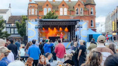 Steven Spicer A pop-up stage is shown in the centre of the picture, with a singer in the middle of the stage wearing a red outfit. Many people are seen gathering to watch the performance.