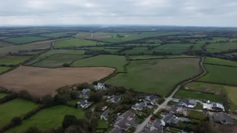 An ariel view of gently rolling green fields and countryside. Part of a hamlet is visable in the foreground, with fields into the distance. One of the fields has a wind turbine.