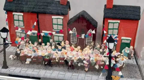 Peterborough Cathedral A knitted display showing a street party celebrating the end of the war. There is a row of three red houses with a brown church in between. In front of them is a very long table with dozens of men, women and children on both sides.