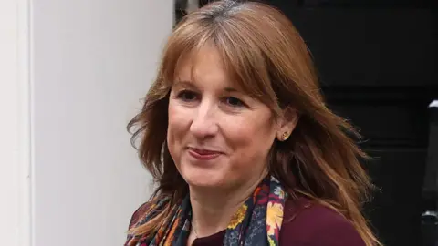 A smiling woman with shoulder-length brown hair is wearing a plum coloured jacket and floral scarf