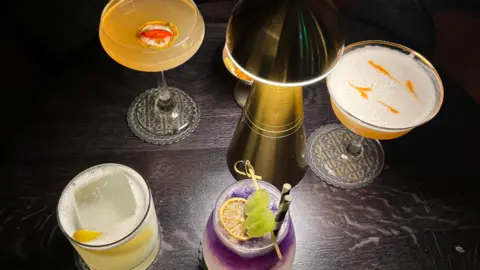 Four cocktails on a dark table with a small lamp on it.