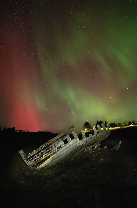 Gary Macleod The Northern Lights in the north coast of Scotland. There are green and red stripes across the sky with the wooden frame of a boat in the foreground. 