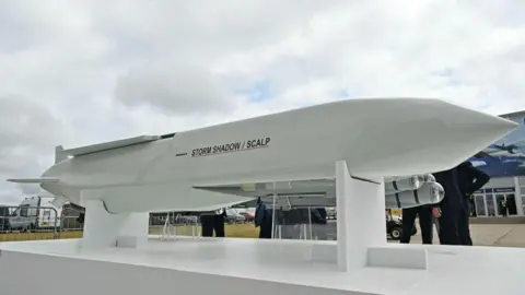 A Storm shadow missile stands against the backdrop of some grey clouds with people wandering around looking at it from the opposite side of the camera.,