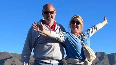 Facebook Andrew and Dawn Searle are smiling for the camera while standing outside - she has a grey jacket/top with her arms stretched out, and a blue top while she is smiling Masu