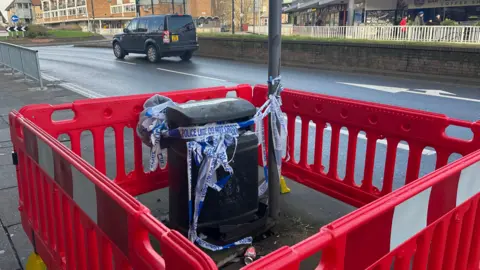 Alice Cunningham/BBC A lamp post and bin are pictured cordoned off with large red plastic fences. Some police tape can be seen wrapped around the lamp post and bin.