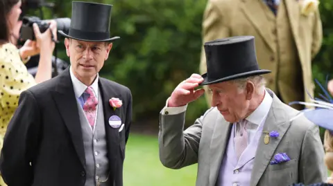 PA Media The King is holding the bill of his top hat and stood next to his brother, Prince Edward. Both are wearing formal suits.